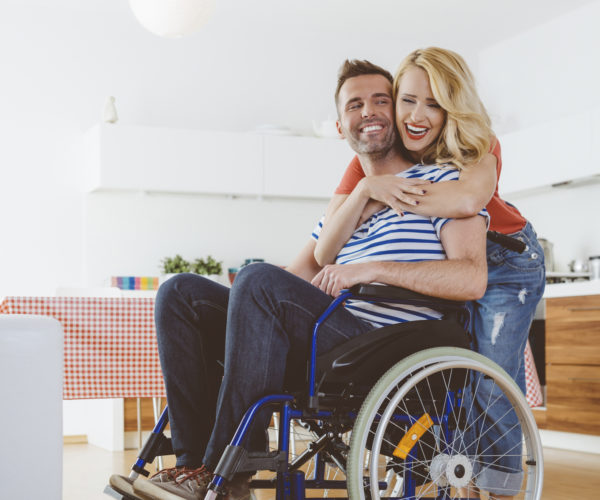 Happy-disabled-man-sitting-in-a-wheelchair-in-the-domestic-kitchen-his-wife-embracing-him