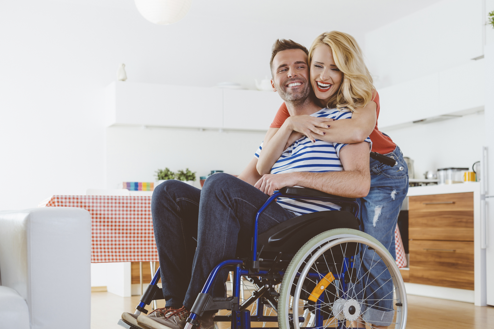 Happy-disabled-man-sitting-in-a-wheelchair-in-the-domestic-kitchen-his-wife-embracing-him